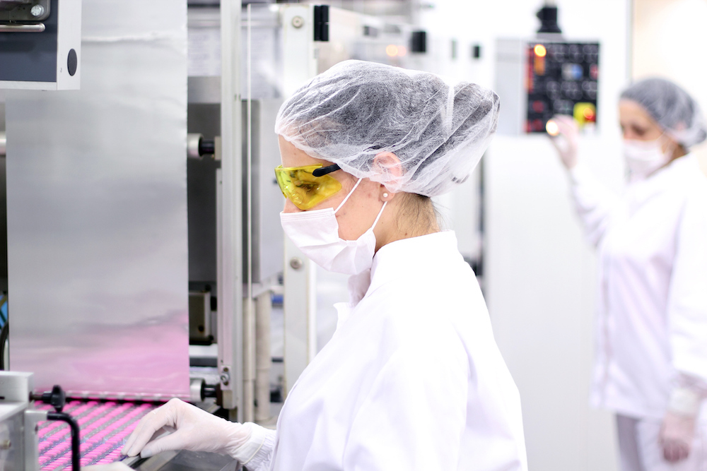 A research in a lab wearing a white hairnet, yellow safety glasses and a white mask and white lab coat is touching a metal shelf with rows of containers with pink lids. Another researcher is in the background.