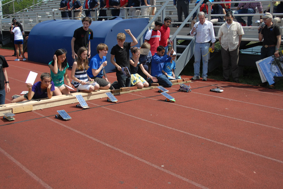 Students racing solar powered cars on an outdoor track as bystanders watch.